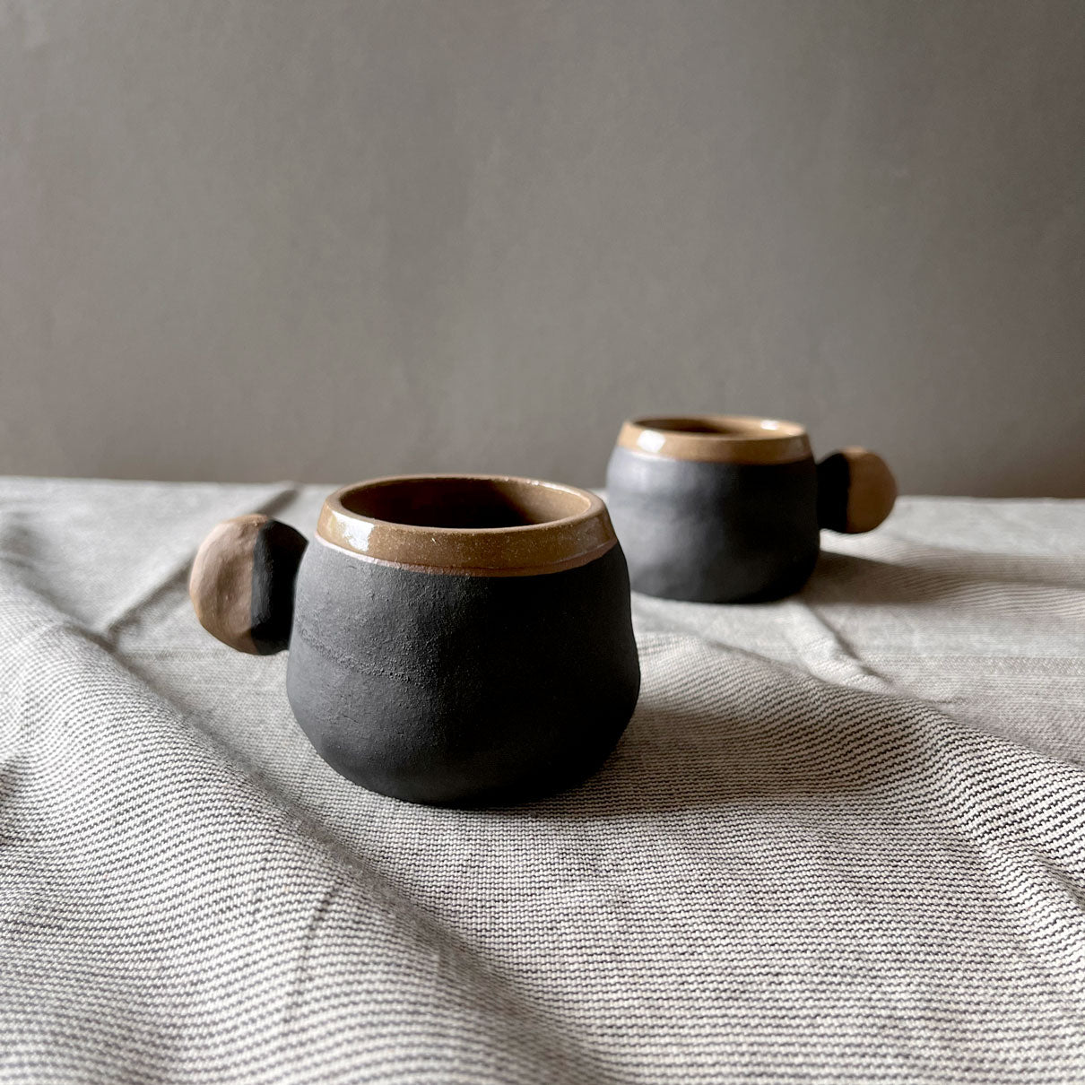 An elegant billowing body form in brown stoneware, featuring a hammered raw texture. Black underglazed exterior that has a volcanic rock texture, with a clear glazed lip and interior.
