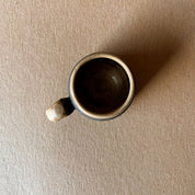 An elegant billowing body form in brown stoneware, featuring a hammered raw texture. Black underglazed exterior that has a volcanic rock texture, with a clear glazed lip and interior.