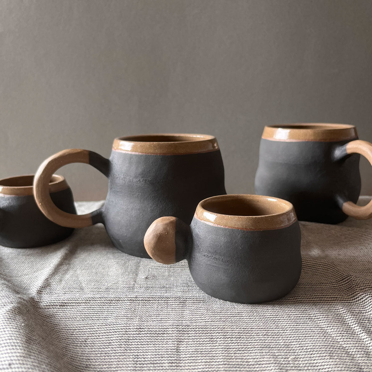 An elegant billowing body form in brown stoneware, featuring a hammered raw texture. Black underglazed exterior that has a volcanic rock texture, with a clear glazed lip and interior.