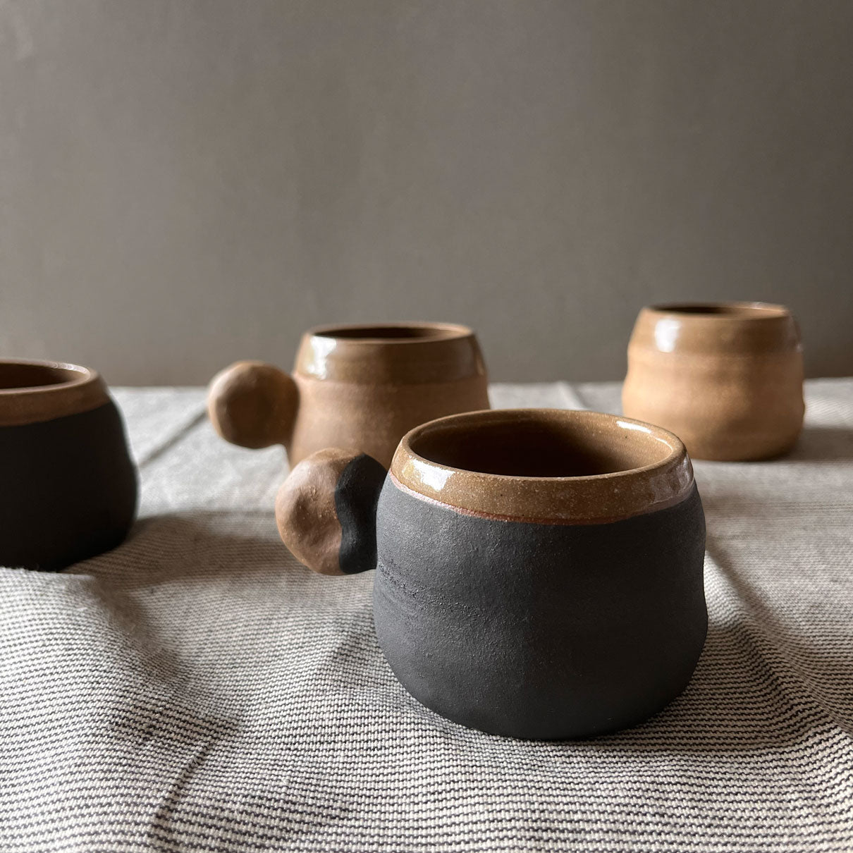 An elegant billowing body form in brown stoneware, featuring a hammered raw texture. Black underglazed exterior that has a volcanic rock texture, with a clear glazed lip and interior.