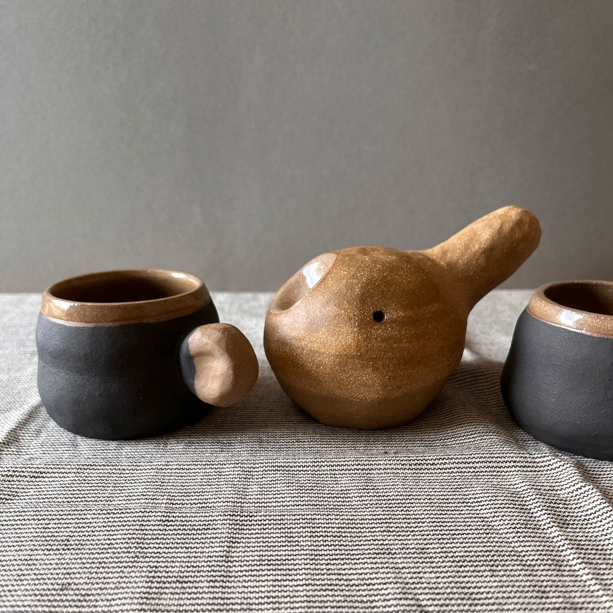 An elegant billowing body form in brown stoneware, featuring a hammered raw texture. Black underglazed exterior that has a volcanic rock texture, with a clear glazed lip and interior.