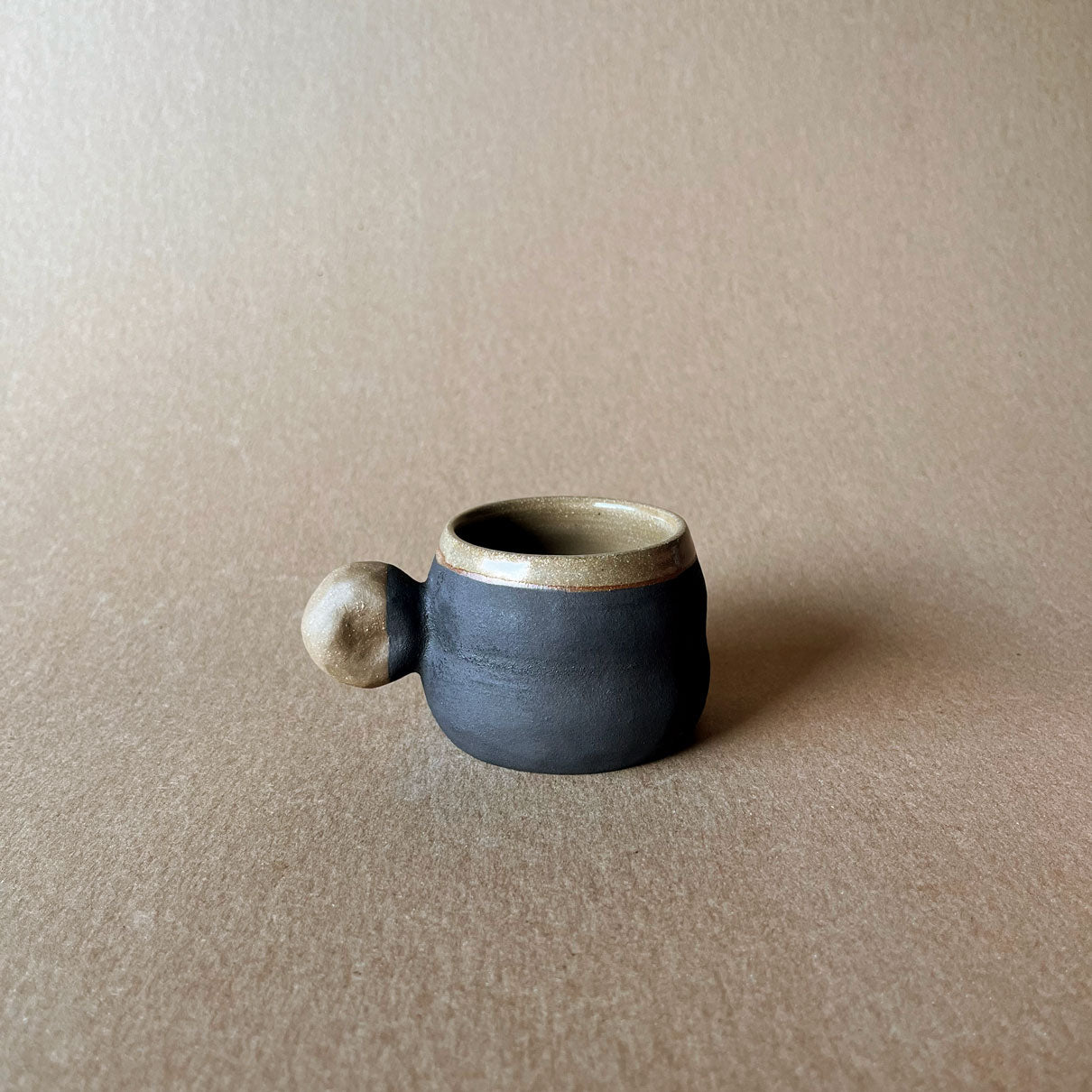 An elegant billowing body form in brown stoneware, featuring a hammered raw texture. Black underglazed exterior that has a volcanic rock texture, with a clear glazed lip and interior.