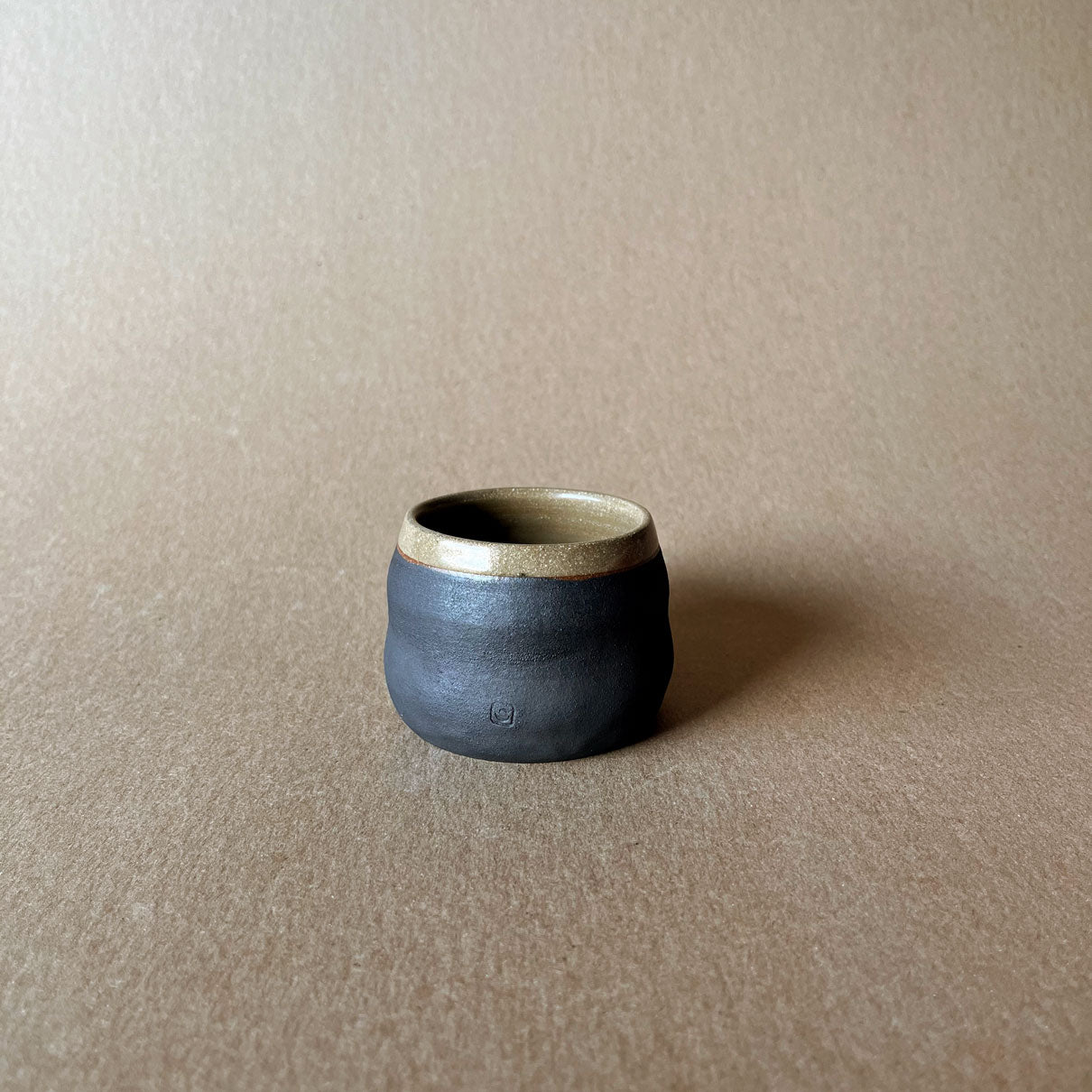 An elegant billowing body form in brown stoneware, featuring a hammered raw texture. Black underglazed exterior that has a volcanic rock texture, with a clear glazed lip and interior.