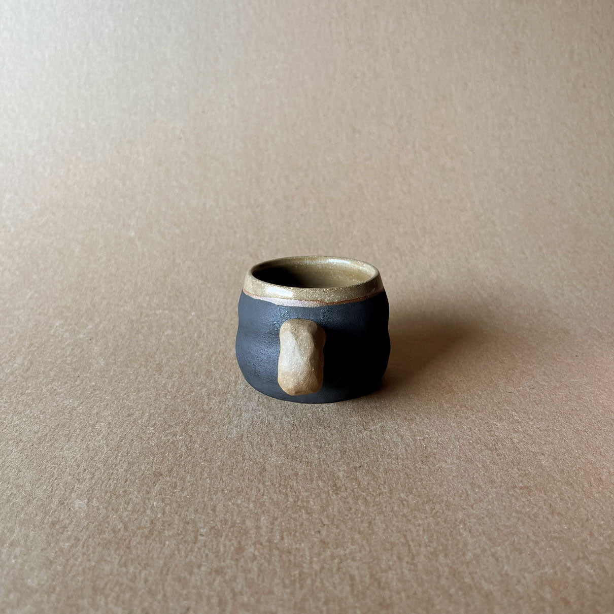 An elegant billowing body form in brown stoneware, featuring a hammered raw texture. Black underglazed exterior that has a volcanic rock texture, with a clear glazed lip and interior.
