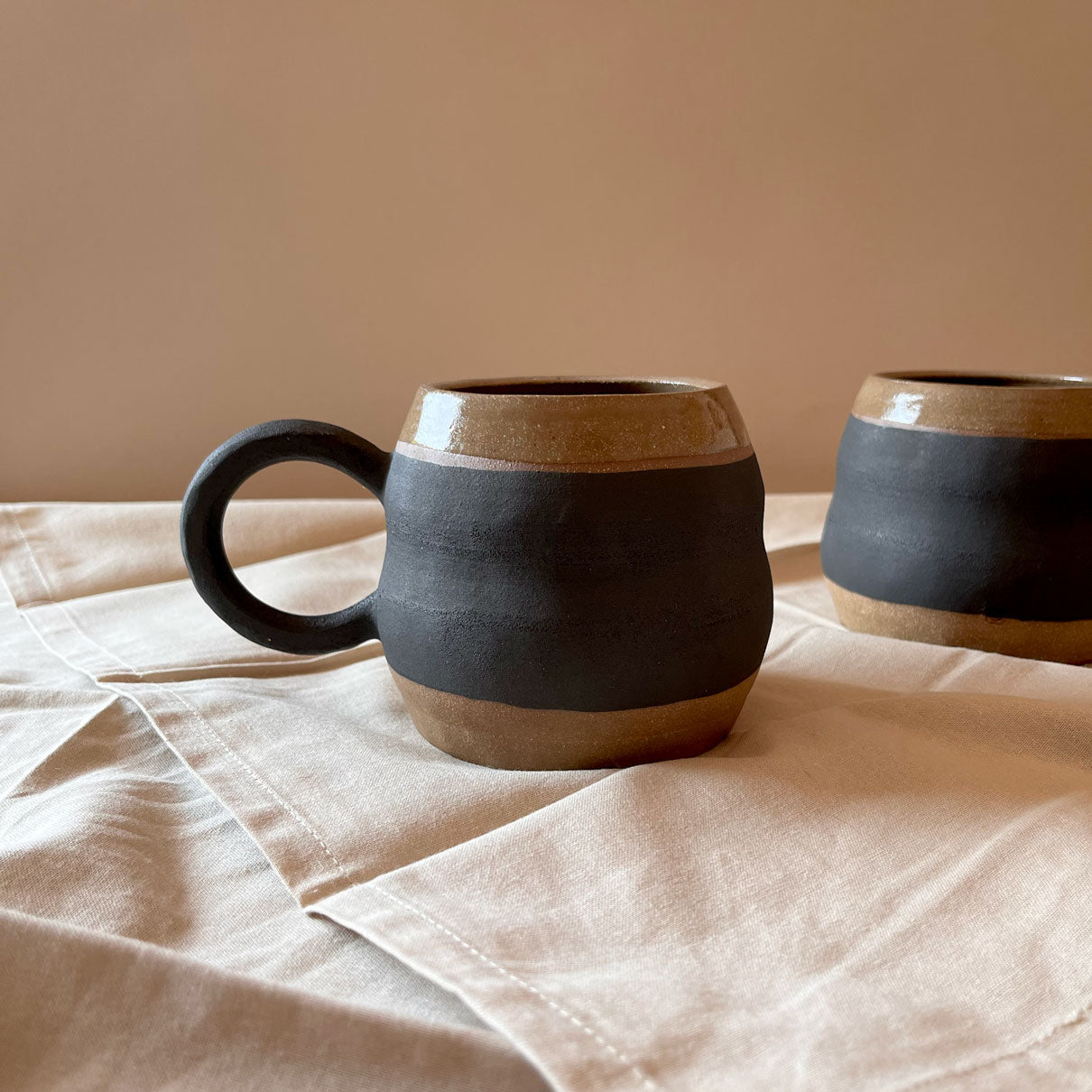 An elegant billowing body form in brown stoneware, featuring a hammered raw texture. Black underglazed exterior that has a volcanic rock texture, with a clear glazed lip and interior.