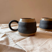 An elegant billowing body form in brown stoneware, featuring a hammered raw texture. Black underglazed exterior that has a volcanic rock texture, with a clear glazed lip and interior.