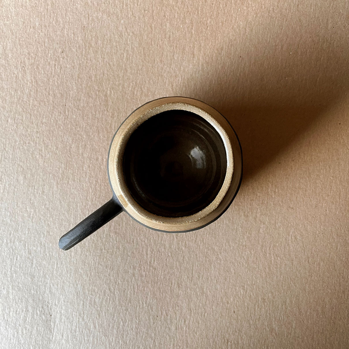 An elegant billowing body form in brown stoneware, featuring a hammered raw texture. Black underglazed exterior that has a volcanic rock texture, with a clear glazed lip and interior.