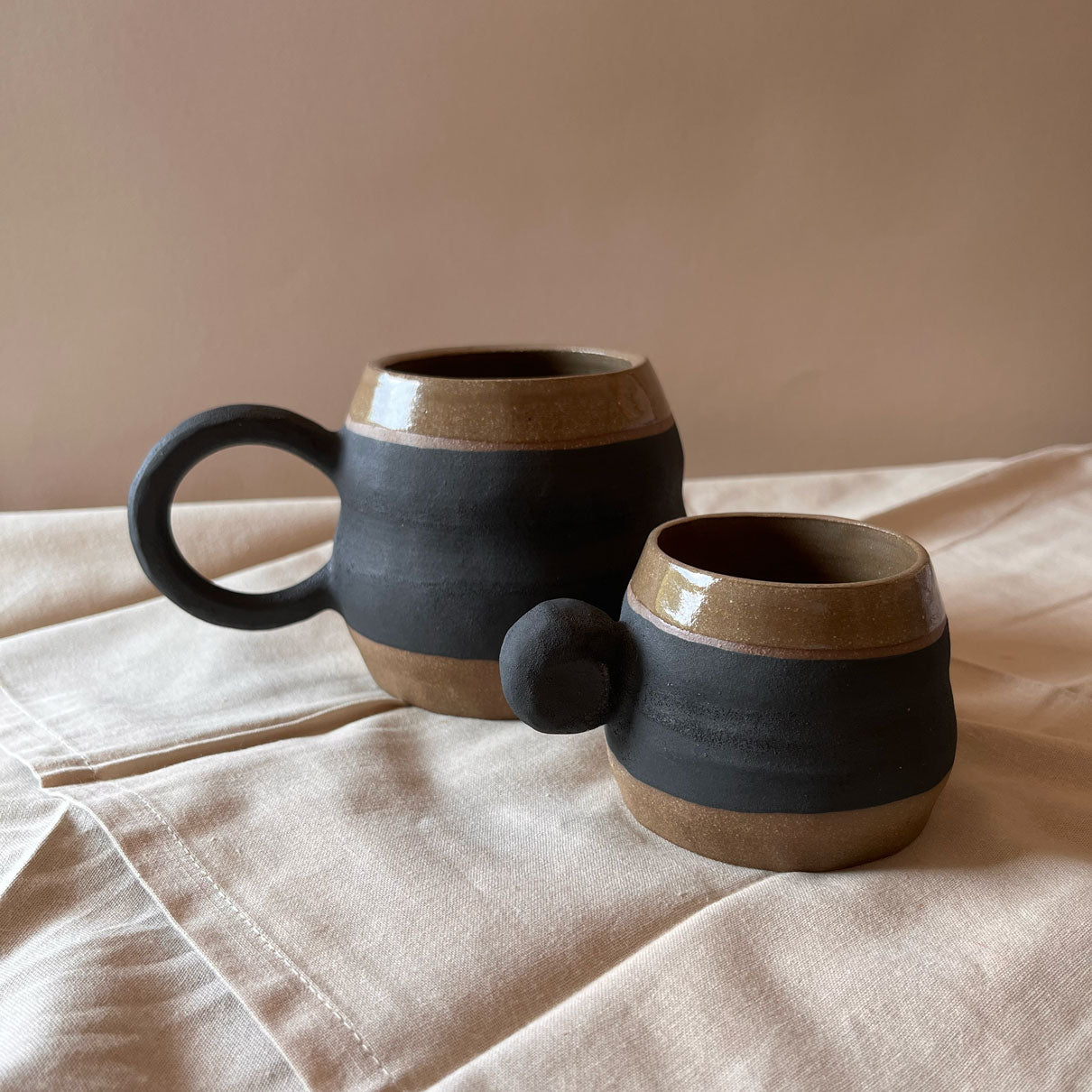 An elegant billowing body form in brown stoneware, featuring a hammered raw texture. Black underglazed exterior that has a volcanic rock texture, with a clear glazed lip and interior.
