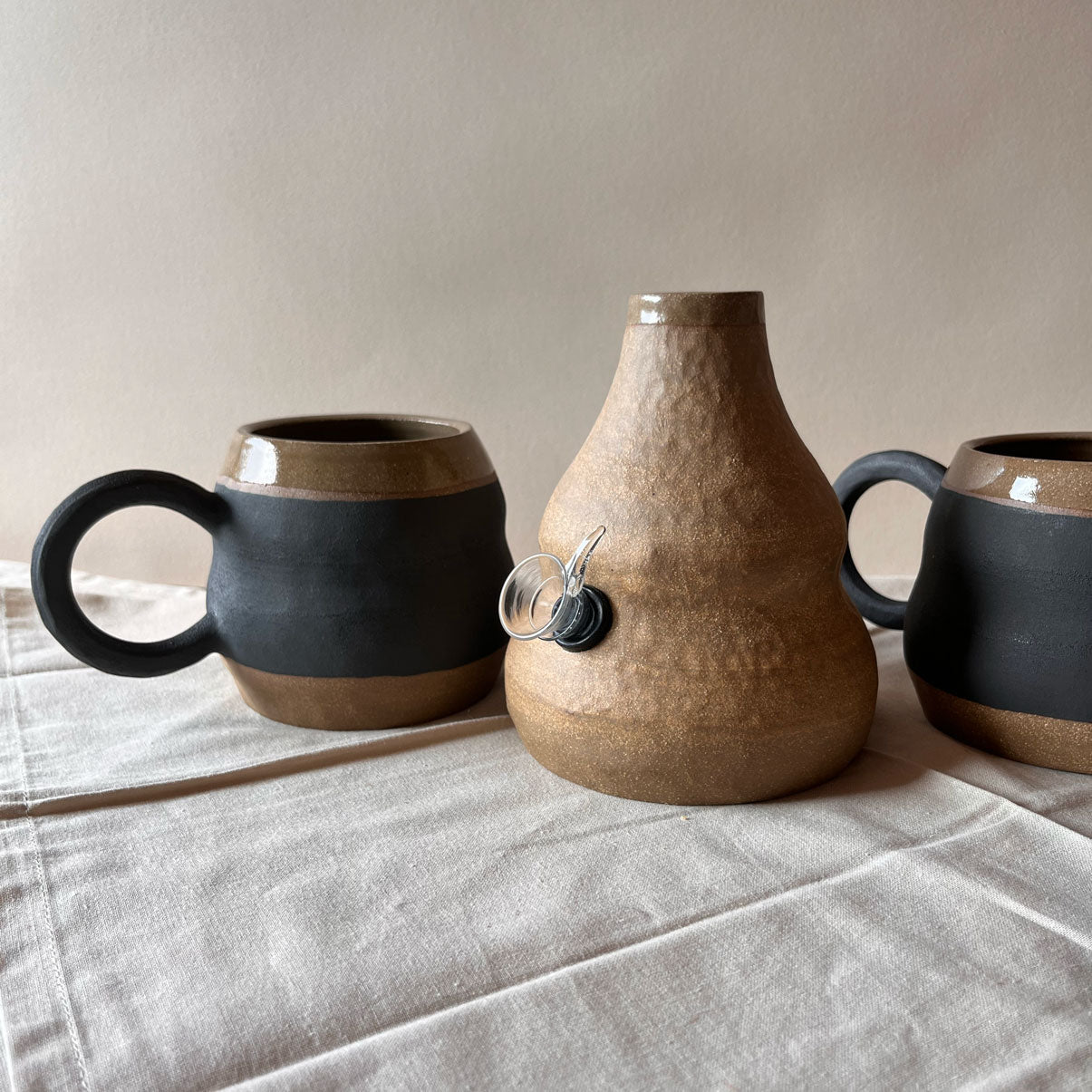 An elegant billowing body form in brown stoneware, featuring a hammered raw texture. Black underglazed exterior that has a volcanic rock texture, with a clear glazed lip and interior.