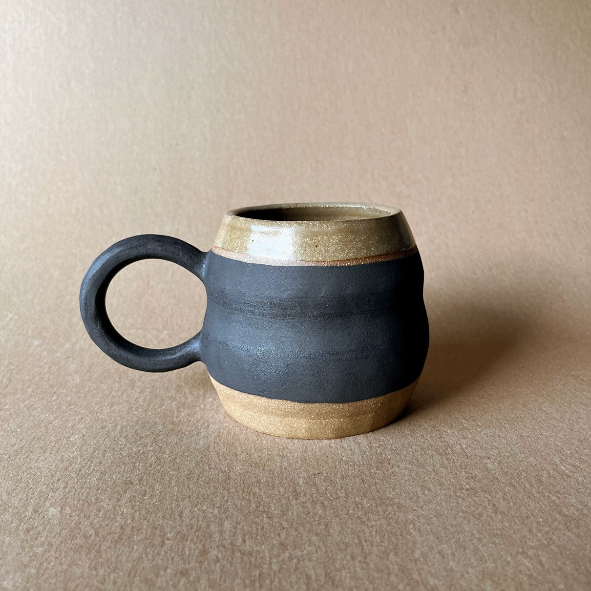 An elegant billowing body form in brown stoneware, featuring a hammered raw texture. Black underglazed exterior that has a volcanic rock texture, with a clear glazed lip and interior.