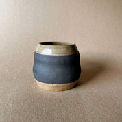 An elegant billowing body form in brown stoneware, featuring a hammered raw texture. Black underglazed exterior that has a volcanic rock texture, with a clear glazed lip and interior.