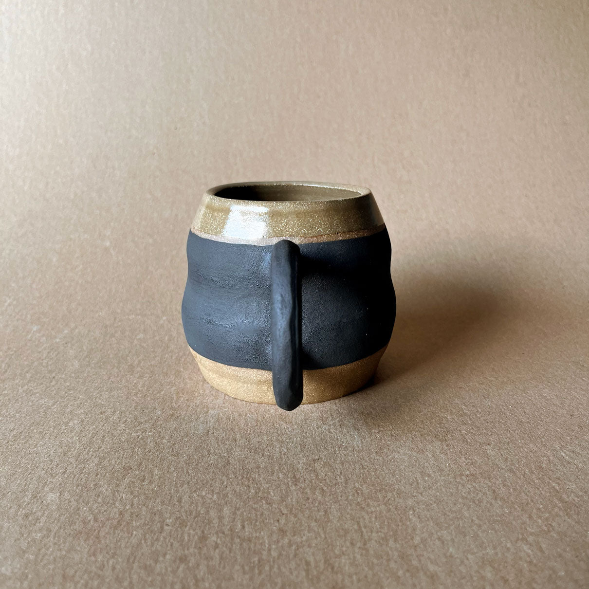 An elegant billowing body form in brown stoneware, featuring a hammered raw texture. Black underglazed exterior that has a volcanic rock texture, with a clear glazed lip and interior.