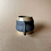 An elegant billowing body form in brown stoneware, featuring a hammered raw texture. Black underglazed exterior that has a volcanic rock texture, with a clear glazed lip and interior.