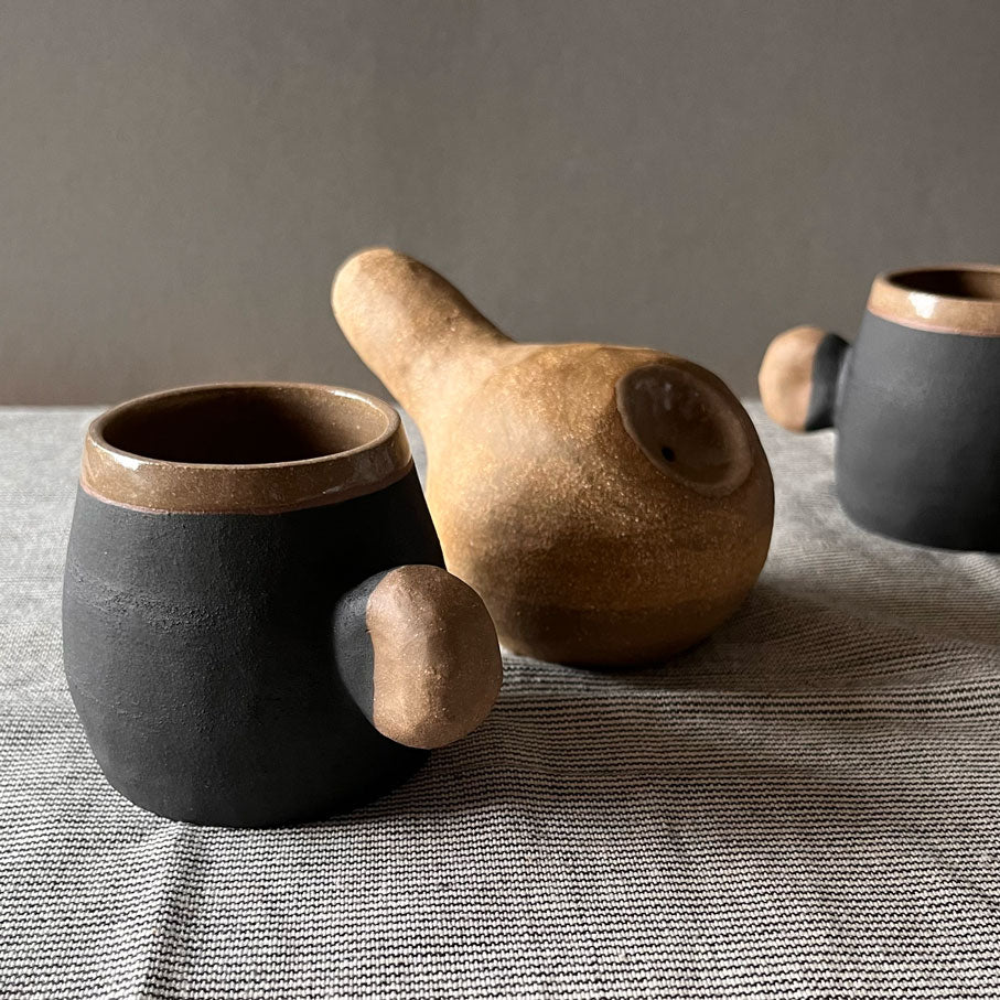 An elegant bulbous body form in brown stoneware, featuring a hammered raw texture. Black underglazed exterior that has a volcanic rock texture, with a clear glazed lip and interior.