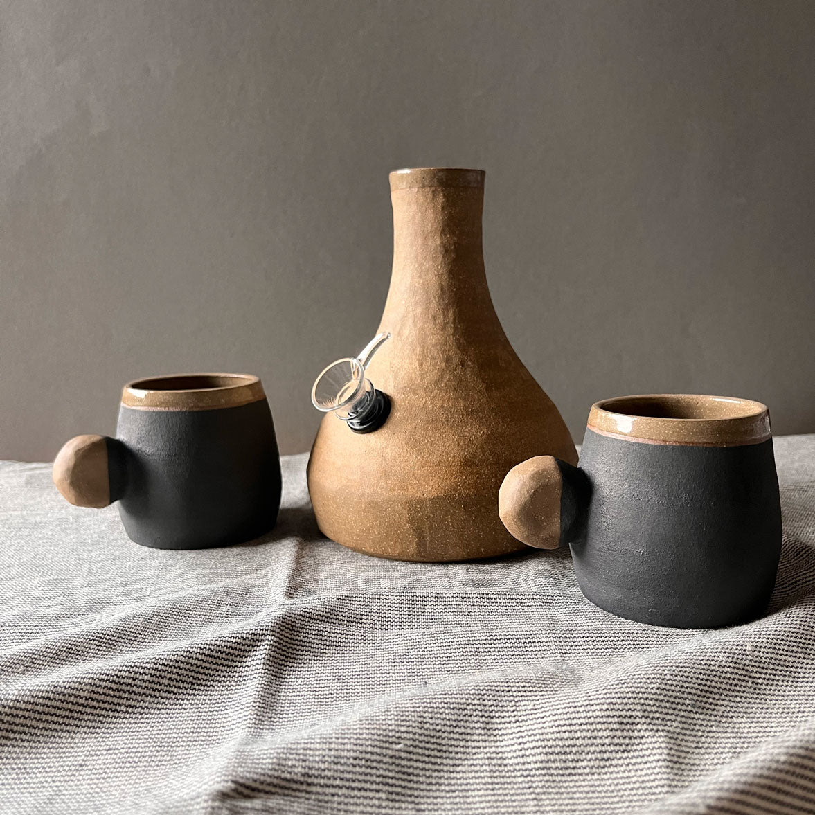 An elegant bulbous body form in brown stoneware, featuring a hammered raw texture. Black underglazed exterior that has a volcanic rock texture, with a clear glazed lip and interior.