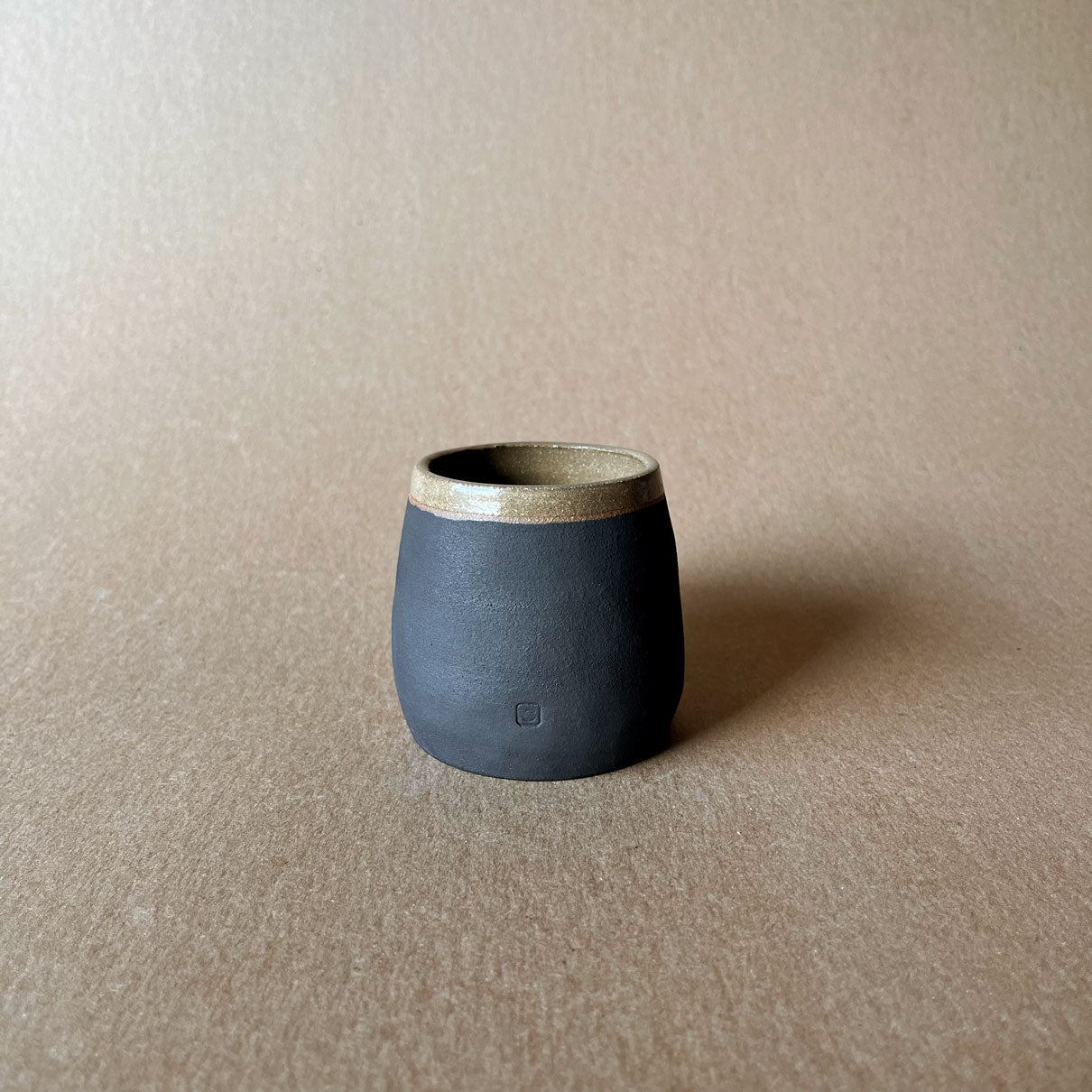 An elegant bulbous body form in brown stoneware, featuring a hammered raw texture. Black underglazed exterior that has a volcanic rock texture, with a clear glazed lip and interior.