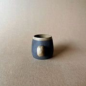 An elegant bulbous body form in brown stoneware, featuring a hammered raw texture. Black underglazed exterior that has a volcanic rock texture, with a clear glazed lip and interior.