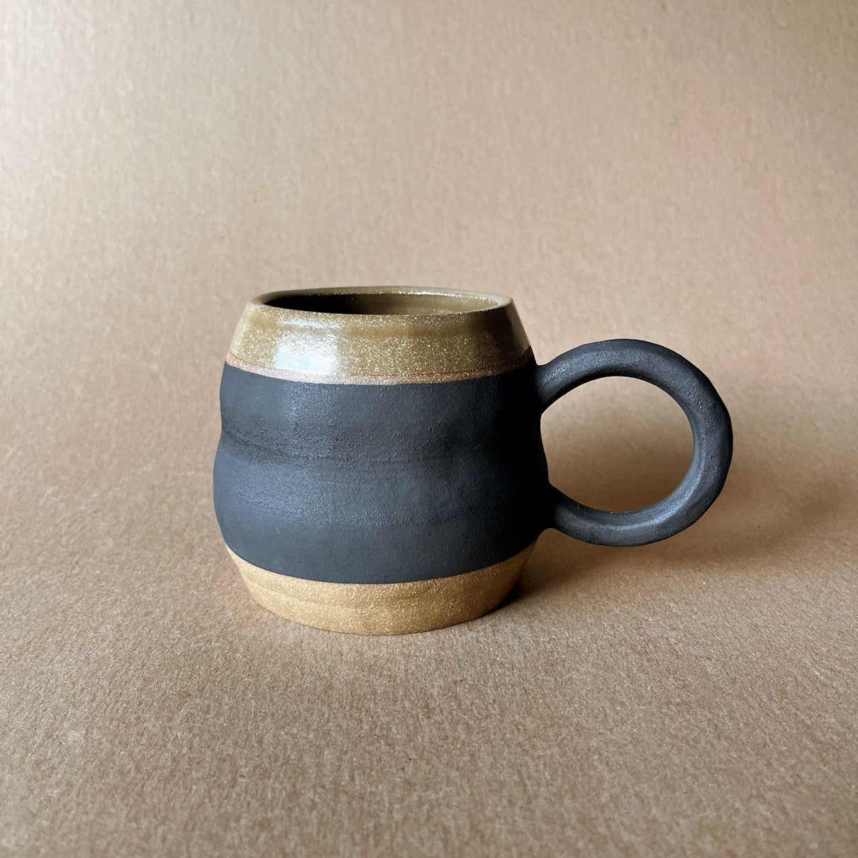 An elegant billowing body form in brown stoneware, featuring a hammered raw texture. Black underglazed exterior that has a volcanic rock texture, with a clear glazed lip and interior. 