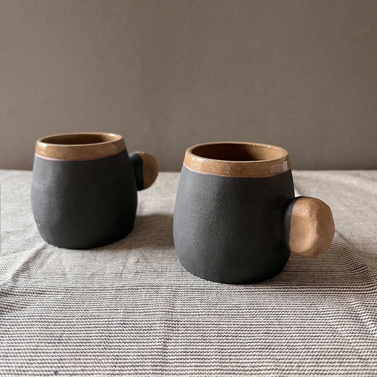 An elegant bulbous body form in brown stoneware, featuring a hammered raw texture. Black underglazed exterior that has a volcanic rock texture, with a clear glazed lip and interior. 