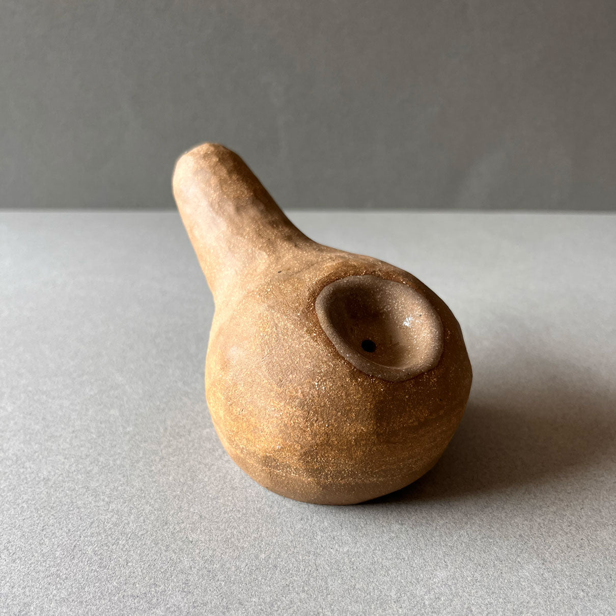 A gourd shaped, spherical body form in brown stoneware with hammered texture, featuring a large glazed inset bowl and rounded stem protrusion mouthpiece. 